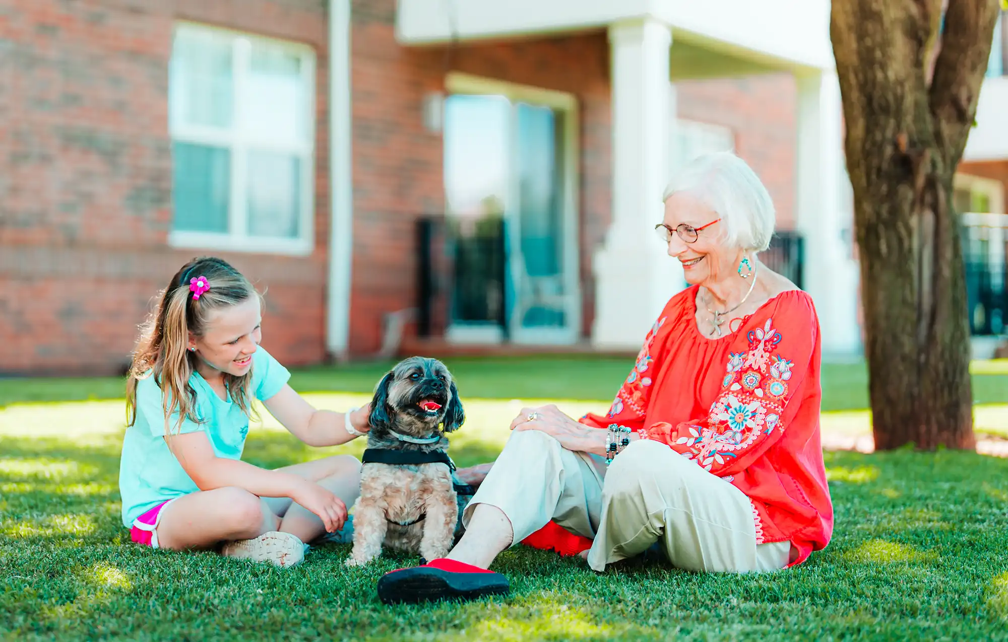 A senior woman and her young granddaughter sit on a lawn with a small dog