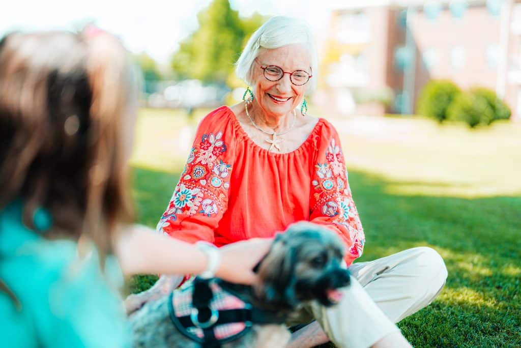 Smiling senior woman in a red embroidered top sits on the lawn petting a small dog