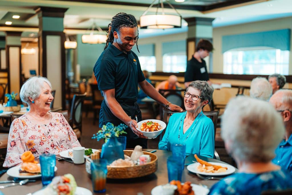 Server delivers plates to smiling senior residents enjoying a meal together